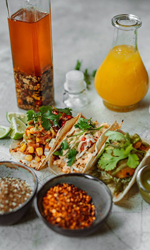 Three tacos filled with corn, chicken, and greens, surrounded by lime wedges, hot sauce, a jar of dressing, and two bowls of spices.