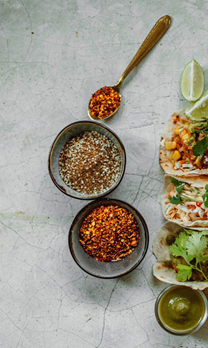 Overhead view of bowls with spices, a spoonful of crushed red pepper, tacos with toppings, lime wedges, and green sauce on a cracked marble surface.