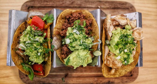 Three tacos arranged on a wooden tray, showcasing their vibrant fillings and garnishes.